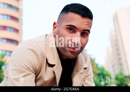 Handsome young unshaven Hispanic male in stylish jacket looking at camera while sitting on modern city street Stock Photo