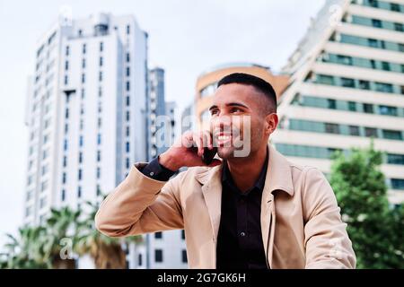 Low angle of positive well dressed young Hispanic businessman talking on smartphone and discussing news on urban street with contemporary buildings in Stock Photo