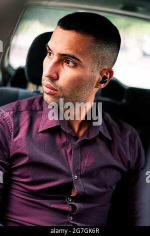 Confident young bearded Hispanic male in stylish shirt using true wireless earbuds while sitting on passenger seat in auto and looking away Stock Photo