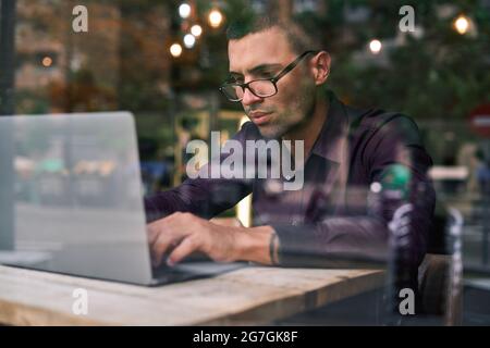 Serious Hispanic male manager in violet shirt and glasses typing on netbook keyboard while sitting at table behind window and working on project in ca Stock Photo