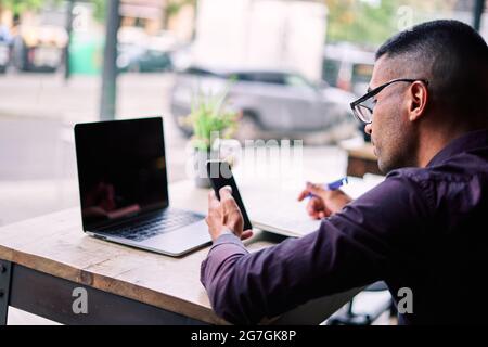 Side view of handsome young Hispanic male entrepreneur checking information on smartphone and writing notes in planner while working at table with lap Stock Photo