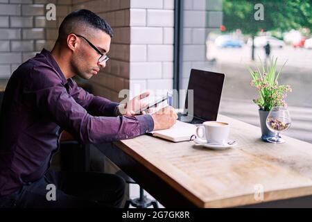 Side view of handsome young Hispanic male entrepreneur checking information on smartphone and writing notes in planner while working at table with lap Stock Photo