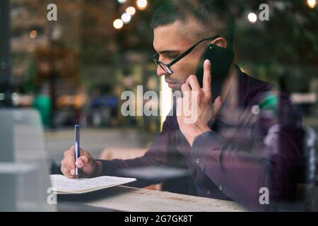 Smart Hispanic businessman in glasses making notes in notepad and answering phone call while sitting behind window in cafeteria Stock Photo