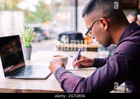 Side view of focused Hispanic man with cup of fresh coffee sitting at table with laptop and making notes in planner while working in cafe Stock Photo