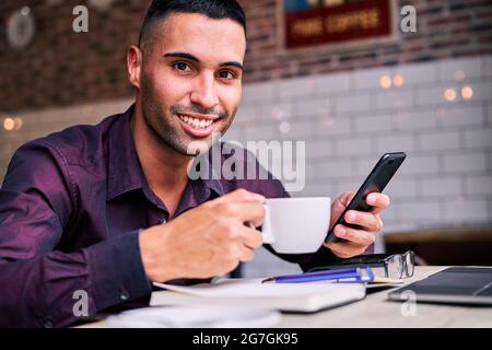 Happy Hispanic male in violet shirt enjoying hot drink and browsing social media on cellphone while taking break during work in cafe Stock Photo