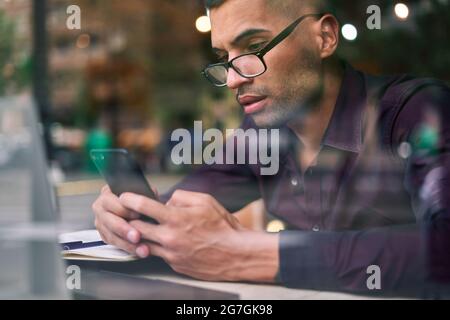 Smart Hispanic businessman in glasses making notes in notepad and answering phone call while sitting behind window in cafeteria Stock Photo