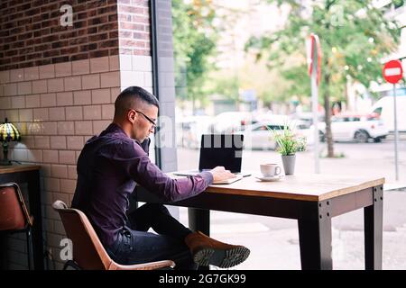 Serious Hispanic male manager in violet shirt and glasses typing on netbook keyboard while sitting at table behind window and working on project in ca Stock Photo