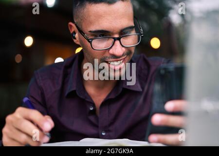 Handsome young Hispanic male entrepreneur checking information on smartphone and writing notes in planner while working at table with laptop Stock Photo