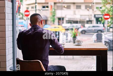 Back view of anonymous male in smart casual clothes sitting at table near window and making notes while working on remote project in cafeteria Stock Photo