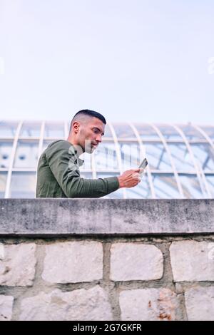 Low angle of adult Hispanic man in shirt leaning on stone wall and browsing cellphone outside contemporary glass building Stock Photo