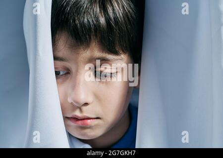 Unhappy little boy peeking out from behind curtains while suffering from domestic violence and hiding from parents Stock Photo