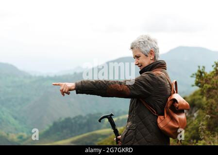 Side view of elderly Woman with backpack and trekking stick standing on grassy slope towards mountain peak during trip in nature pointing with finger Stock Photo