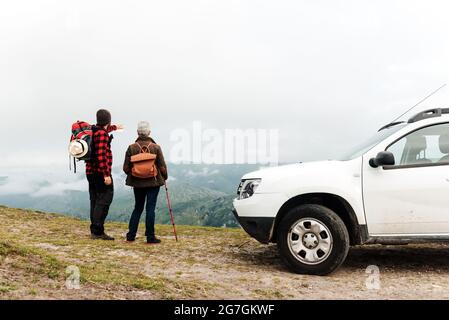 Senior Woman and adult man discussing route together while standing near white vehicle during road trip in mountains Stock Photo