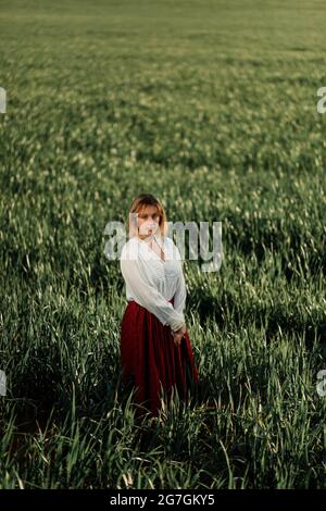 Calm young female dressed in old fashioned blouse and skirt standing alone among tall green grass in cloudy summer day in countryside Stock Photo
