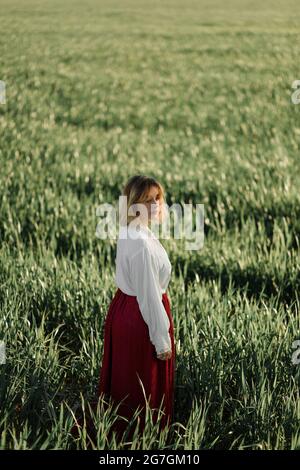Calm young female dressed in old fashioned blouse and skirt standing alone among tall green grass in cloudy summer day in countryside Stock Photo