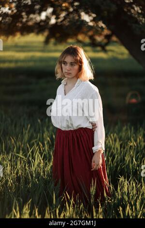 Calm young female dressed in old fashioned blouse and skirt standing alone among tall green grass in cloudy summer day in countryside Stock Photo