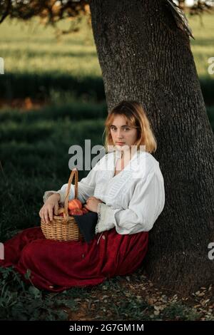 Young female in old fashioned white blouse and skirt holding wicker basket full of fresh apples and looking at camera while resting near tree in summe Stock Photo