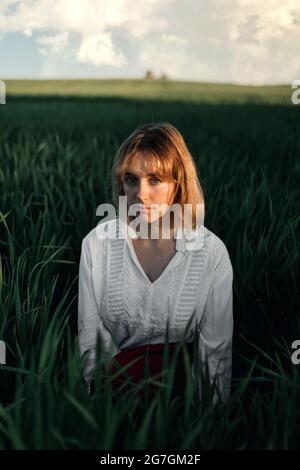 Peaceful young female in retro styled white blouse sitting amidst tall green grass and looking at camera while resting in summer evening in countrysid Stock Photo