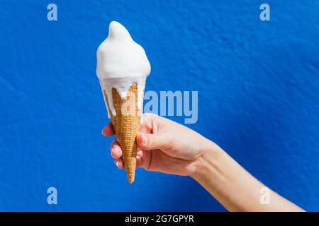 Crop hand of anonymous female with melting ice cream in waffle cone on sunny day on blue background Stock Photo