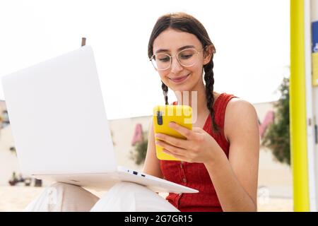 Content female freelancer sitting on seashore and typing on laptop and browsing on smartphone while working remotely on project in summer Stock Photo