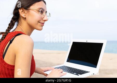 Content female freelancer sitting on seashore and typing on laptop while working remotely on project in summer Stock Photo