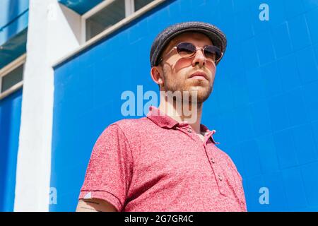 Young bearded guy wearing casual red polo shirt and cap standing against blue wall on street in summer day Stock Photo