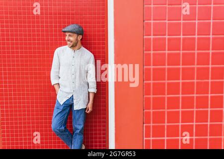 Handsome young bearded guy in trendy casual shirt and jeans and cap standing near red wall and looking away with smile Stock Photo