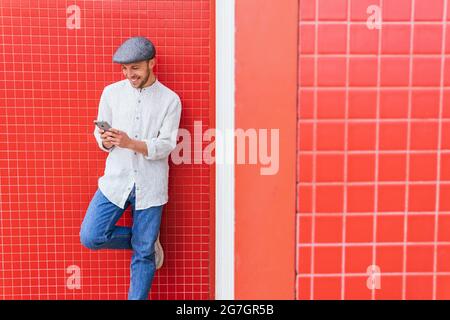 Content young bearded hipster guy in casual polo shirt and cap browsing mobile phone while standing against red wall in sunlight Stock Photo