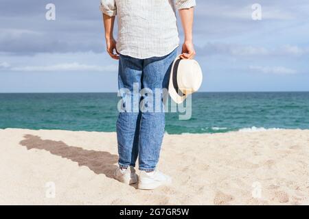 Back view of cropped unrecognizable male in trendy casual clothes and hat walking alone on sandy beach towards waving sea while spending summer holida Stock Photo