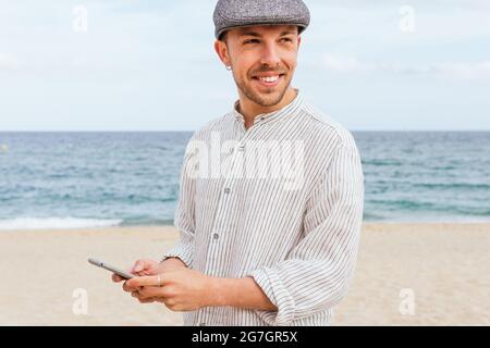 Positive young bearded guy in stylish shirt and cap smiling and looking away while browsing mobile phone on sandy beach near sea Stock Photo