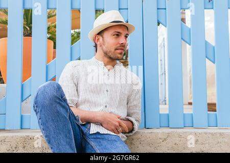 Handsome young bearded guy in stylish striped shirt and straw hat looking away while chilling near blue fence in summer day Stock Photo