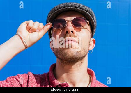Young bearded guy wearing casual red polo shirt and cap standing against blue wall on street in summer day Stock Photo