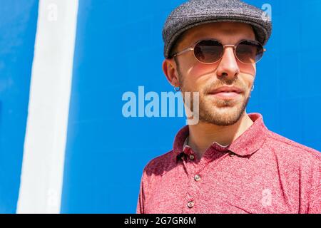 Young bearded guy wearing casual red polo shirt and cap standing against blue wall on street in summer day Stock Photo