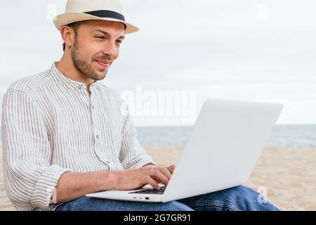 Side view of young male blogger in stylish wear and hat sitting on sandy beach and typing on laptop during summer holidays on seashore Stock Photo