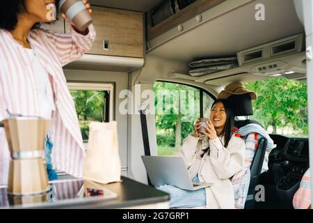 Cheerful young Asian Woman with laptop drinking coffee with unrecognizable friend inside camper van during summer journey in nature Stock Photo