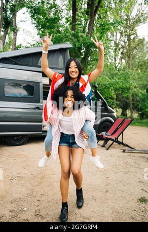 Full length of cheerful young African American Woman giving piggyback ride to content Asian girlfriend with American flag while enjoying summer advent Stock Photo
