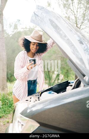 Young African American female traveler holding smartphone with glowing flashlight while checking car engine as having problem during road trip through Stock Photo