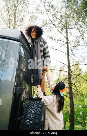 Side view of young African American Woman helping friend climbing up camper van parked in green forest during summer adventure in countryside Stock Photo