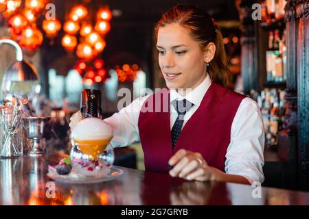 Skilled young female barkeeper using flavor bluster smoke gun while garnishing cocktail at bar counter Stock Photo