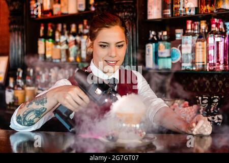 Skilled young female barkeeper using flavor bluster smoke gun while garnishing cocktail at bar counter Stock Photo