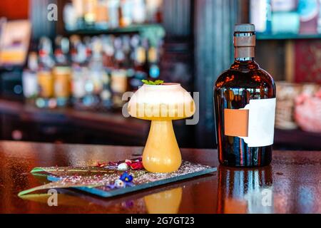 Sour cocktail with mint leaf in mushroom shaped glass served on wooden counter with bottle of alcohol drink in pub Stock Photo