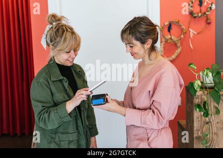 Smiling female client making contactless payment on POS terminal with mobile phone while standing in spa salon Stock Photo