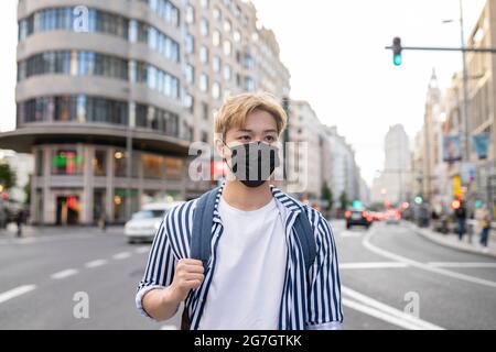Ethnic male student with backpack and in protective mask standing in street during coronavirus epidemic Stock Photo