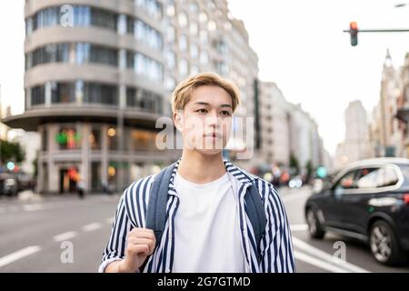 Trendy Asian male model with blond hair and backpack standing in city street and looking away Stock Photo