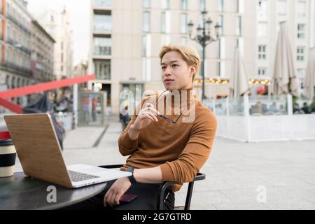 Pensive Asian male freelancer sitting with laptop at table in street cafe and working remotely on startup while looking away Stock Photo