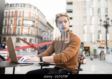 Pensive Asian male freelancer sitting with laptop at table in street cafe and working remotely on startup while looking away Stock Photo