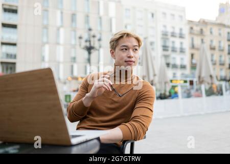 Pensive Asian male freelancer sitting with laptop at table in street cafe and working remotely on startup while looking away Stock Photo