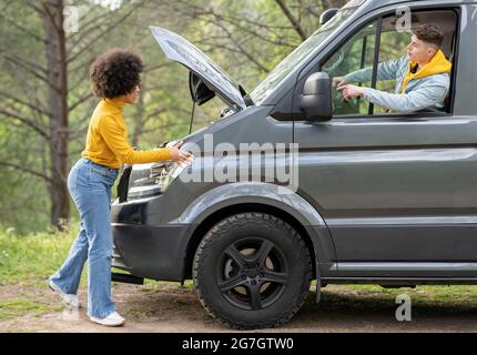Side view of black Woman with Afro hairstyle listening to boyfriend instructions and checking van engine while trying to repair vehicle during trip th Stock Photo