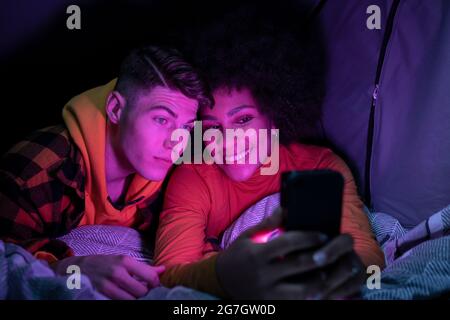Happy multiracial man and Woman smiling resting and browsing cellphone in tent at night Stock Photo