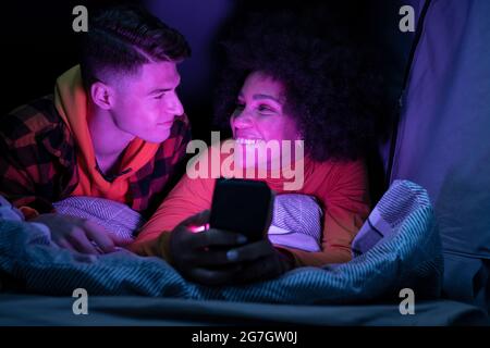 Happy multiracial man and Woman smiling and looking at each other while resting and browsing cellphone in tent at night Stock Photo
